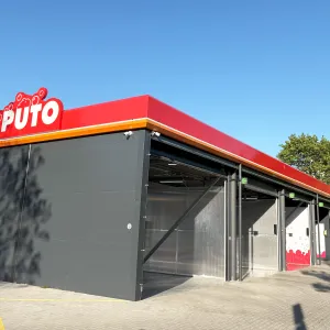 Pelēks automazgātavas ēka ar sarkanu jumtu skaidrās debesīs.