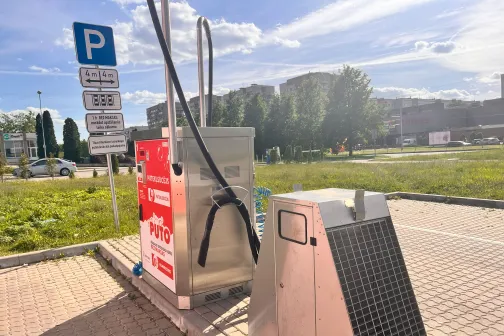 Autostāvvieta saulainā dienā ar iekārtām, zālienu un kokiem fonā.