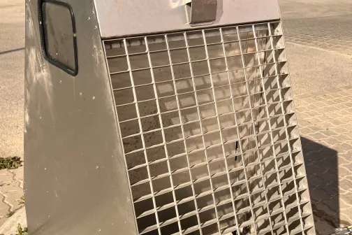 Metāla konteineris ar režģi sānā uz ietves saulainā dienā.