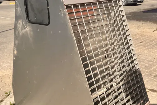 Metāla režģa konstrukcija uz ietves, fonā krāsainas ēkas.