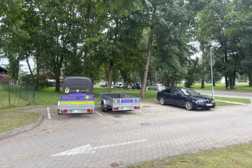 Stāvvietā zem kokiem stāv divas automa&scaron;īnas pie piekabēm.