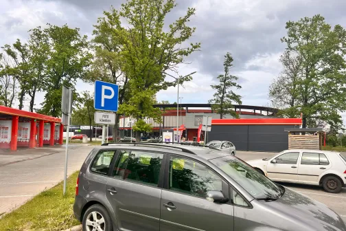 Autostāvvieta pie sarkanām ēkām un kokiem, mākoņains laiks.