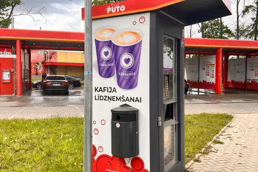 Kafijas automāts stāvlaukumā pie sarkanas nojumes.