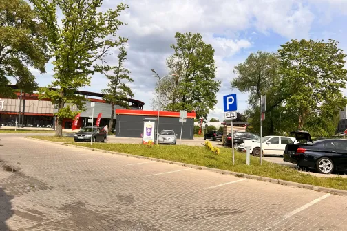 Autostāvvieta ar kokiem, debesīs mākoņi, centrā stāvvietas zīme.