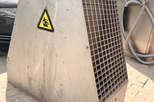 Metāla skapis ar režģi un brīdinājuma zīmi uz ietves.