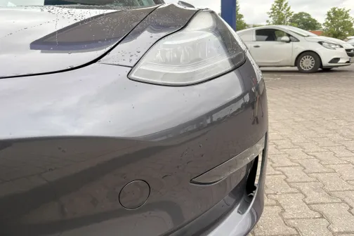 Tum&scaron;as automa&scaron;īnas priek&scaron;puse ar ūdens pilieniem, fonā stāv balta automa&scaron;īna.