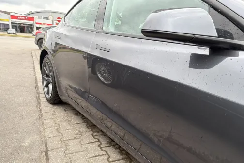 Melns auto stāvvietā, blāvi atspīdumi sānos.