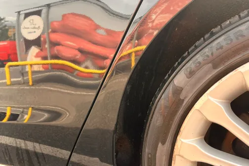 Auto sānu atspīdums ar spilgtiem objektiem fonā, redzams detaļas un riepa.