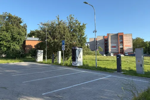 Tuk&scaron;a autostāvvieta ar zaļiem kokiem un ēkām fonā, dzidras debesis.