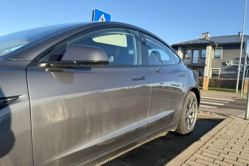 Netīrs sudraba auto saulainā ielā pie ietves.