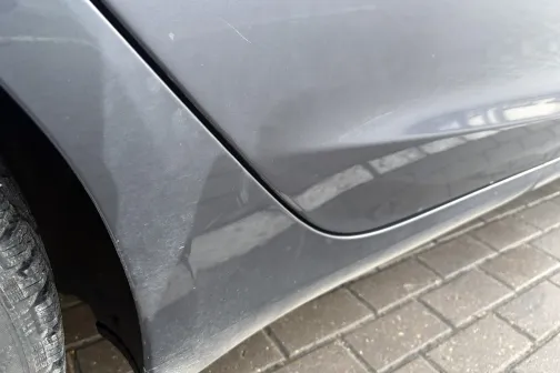 Netīrs pelēks auto durvju panelis, blakus riepa, bruģēts segums.