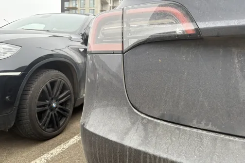 Divi netīri automobiļi stāv stāvvietā blakus ēkai.