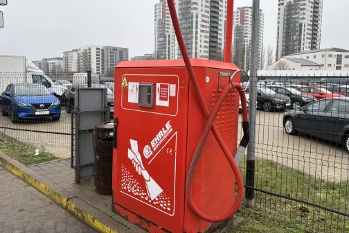 Sarkans putekļu sūcējs autostāvvietā ar daudzstāvu mājām fonā.