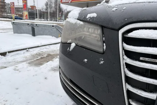 Melnas automa&scaron;īnas priek&scaron;ējais lukturis ar sniegu, fonā redzama auto mazgātava.