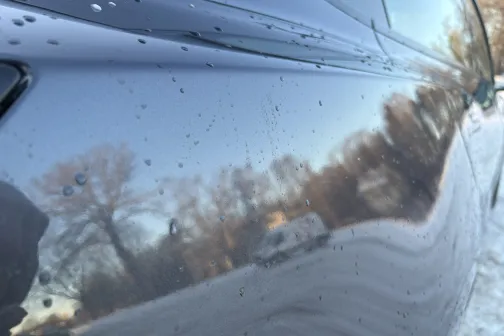 Auto netīra virsma ar ūdens pilieniem un koku atspulgiem.