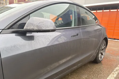 Netīrs pelēks automobilis stāvvietā blakus oranžai sienai.