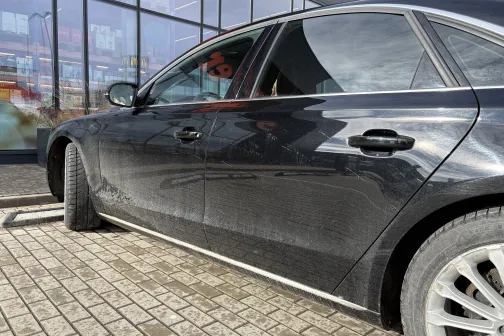 Melns auto ar dubļiem uz sāniem, novietots pie stiklotas ēkas.