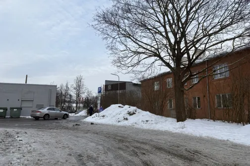 Ziema ārā, ceļ&scaron; ar sniegu un kailiem kokiem, pelēka ēka fonā.