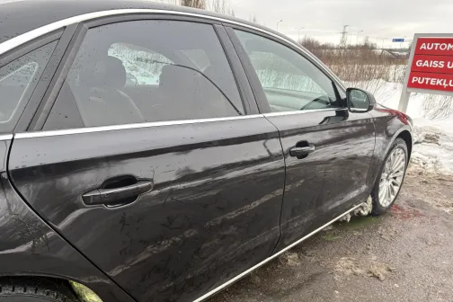 Melns automobilis ar netīriem sāniem, ceļa malā, redzama reklāmas zīme.