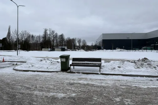 Sniegots stāvlaukums ar soliņu un atkritumu tvertni pie ēkas.