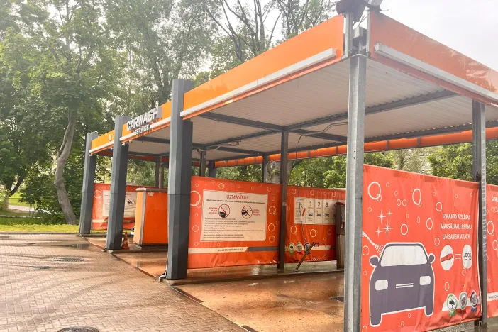 Automa&scaron;īnu mazgātava ar oranžiem un sarkaniem rotājumiem, zaļi koki fonā.