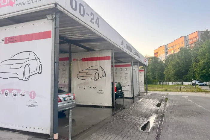 Āra automazgātava ar auto zīmējumiem, blakus daudzstāvu ēka un zaļi koki.