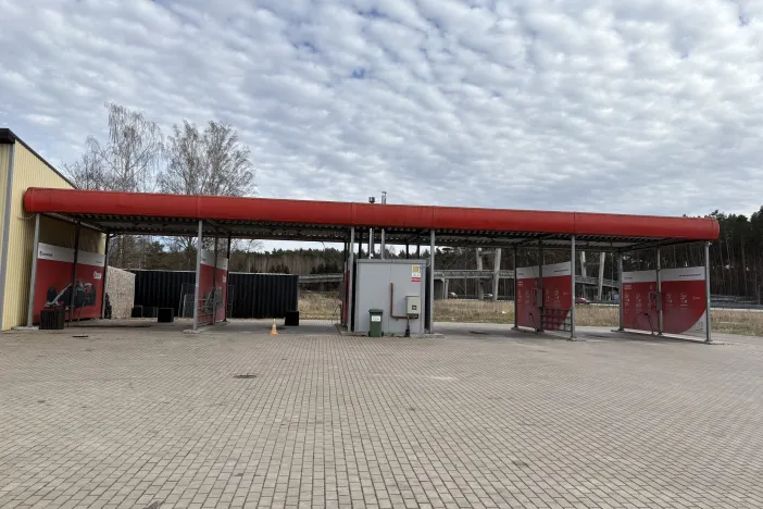 Āra automazgātuve ar sarkanām un pelēkām konstrukcijām, mākoņainas debesis.